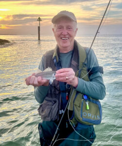 Flyfisherman at dawn on the dorset coast with sun behind clouds behind, fisherman in cap with rod holding a sea bass