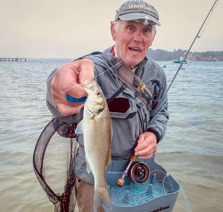 angler holding sea bass up and fly fishing rod in left hand with sea in background