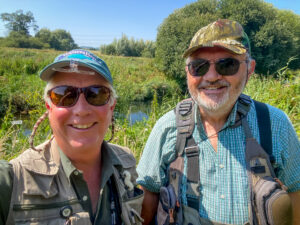 2 happy fly fishing anglers on the Dorset Allen with trees, reeds and river, and blue sky behind