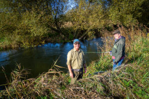 2 fishermen by river one holding trotting rod