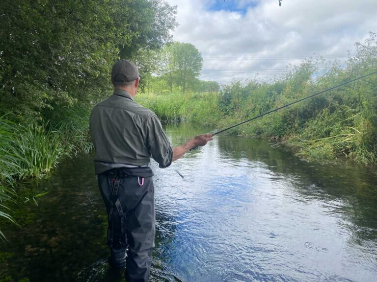 man flyfishing a chalkstream with sky and bankside vegetation